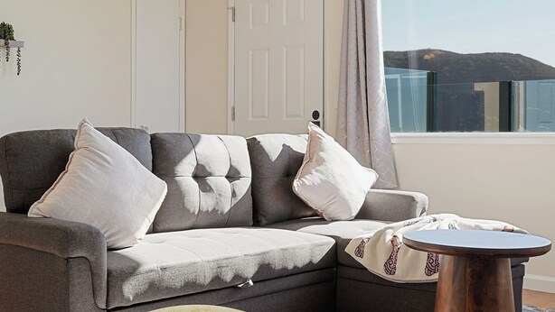 Ein modernes Wohnzimmer mit einem grauen Ecksofa, einem runden Couchtisch aus Holz und einem senffarbenen Hocker.