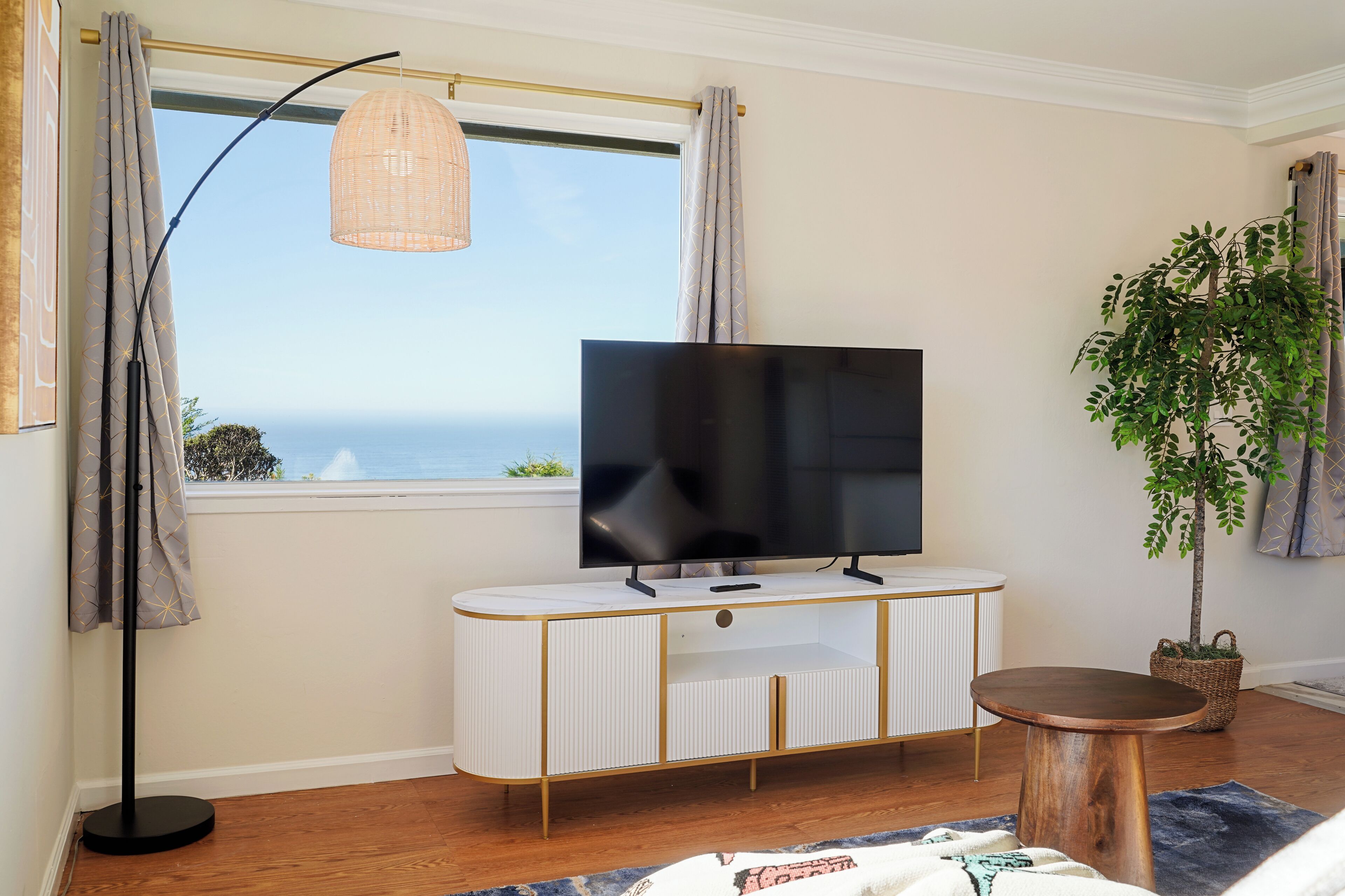 En moderne stue med et stort fladskærms-tv på et hvidt og gyldent konsolbord, en gulvlampe, et rundt træbord og en potteplante.