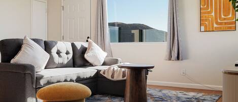 A living room with a sofa, ottoman, and round table, a large window with curtains, and a wall-mounted artwork.