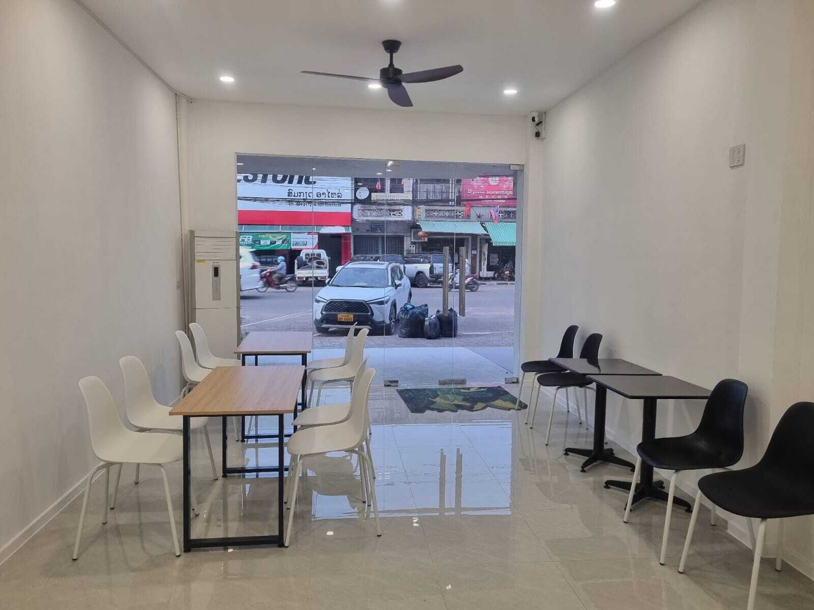 Uma sala de conferências com mesas, cadeiras, um ventilador de teto e vista para a rua.