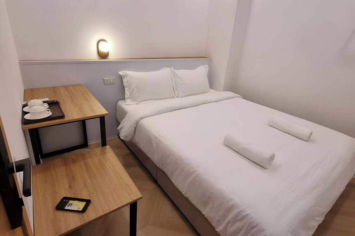 Une chambre d’hôtel bien rangée, avec un lit, une table de chevet sur laquelle se trouve une assiette remplie de tasses blanches, une petite table avec une tablette, et un lampadaire fixé au mur.
