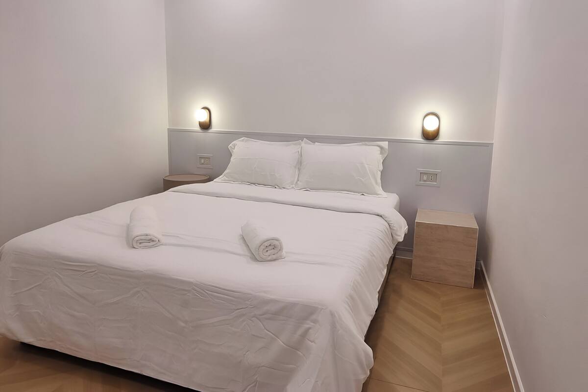 Un lit bien fait, avec du linge de lit et des serviettes blanches, deux tables de chevet et des lampes fixées au mur.