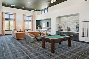 A modern living room with a pool table, a sofa set, and a kitchen area with a brick backsplash.