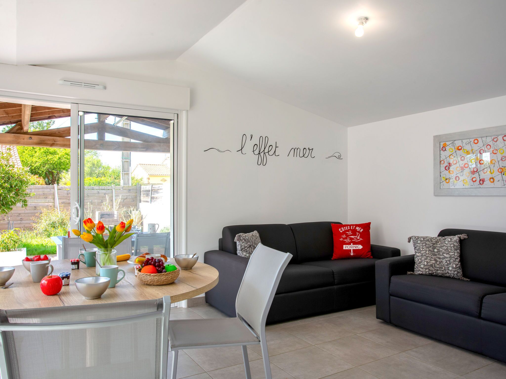 Een moderne woonkamer met een zwarte bank, een eettafel met fruit en een muur waarop een Franse uitdrukking staat.