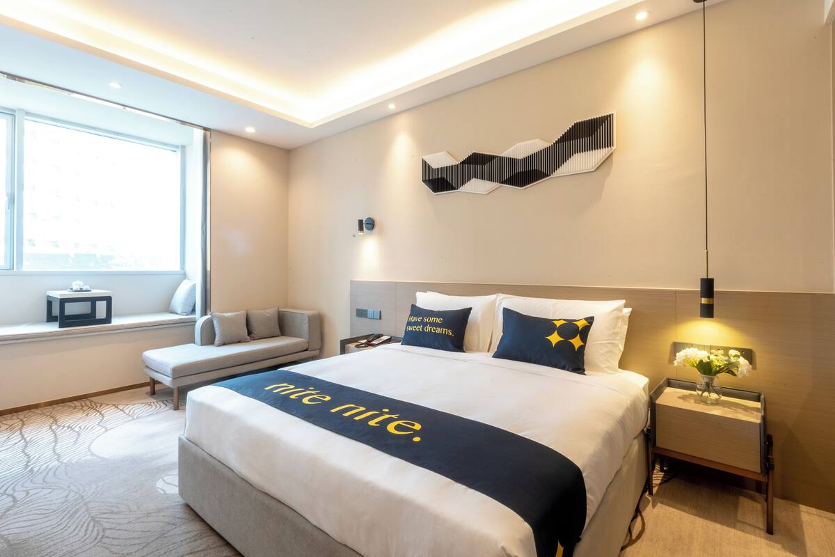 Une chambre d’hôtel avec un grand lit, une table de chevet avec une lampe, une fenêtre donnant sur un beau paysage et une œuvre d’art accrochée au mur.