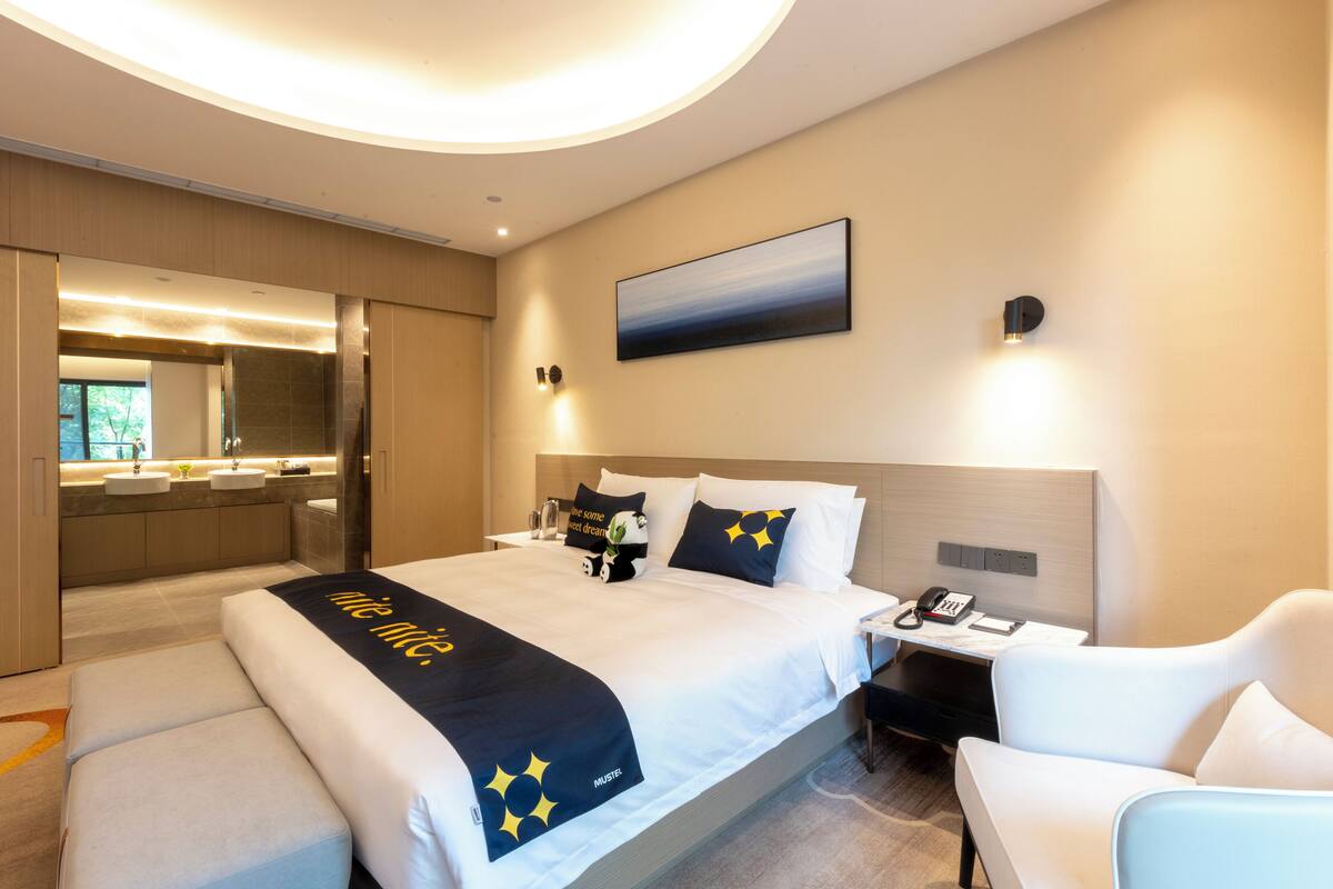Une chambre d’hôtel moderne dotée d’un grand lit, d’une table de chevet, d’une chaise, d’un bureau, d’un miroir et d’une salle de bain avec un lavabo et une douche.