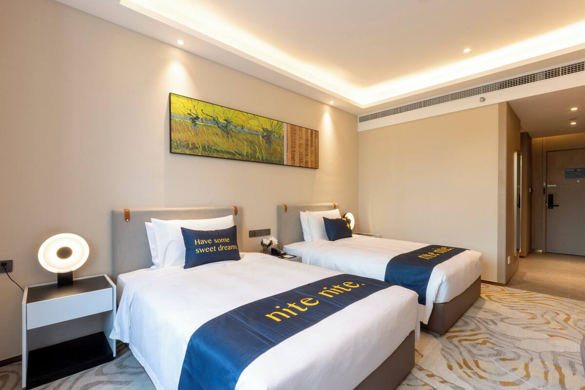 Une chambre d’hôtel avec deux lits, une table de chevet avec une lampe, un tableau au mur et un sol recouvert de moquette.