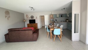 A modern living room with a brown sofa, a dining table with chairs, a TV on a wooden cabinet, and a kitchen area with cabinets and appliances. - Cocooning villa with jacuzzi (Le Grau-du-Roi)
