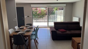 A dining area with a table set for four, a sofa, and a sliding glass door leading to a balcony with a view of trees and a railing. - Cocooning villa with jacuzzi (Le Grau-du-Roi)