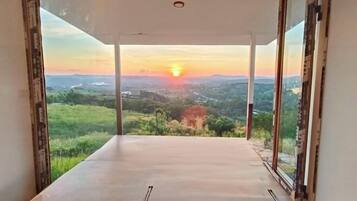 A room with a large glass door leading to a balcony with a scenic view of a sunset over a valley.