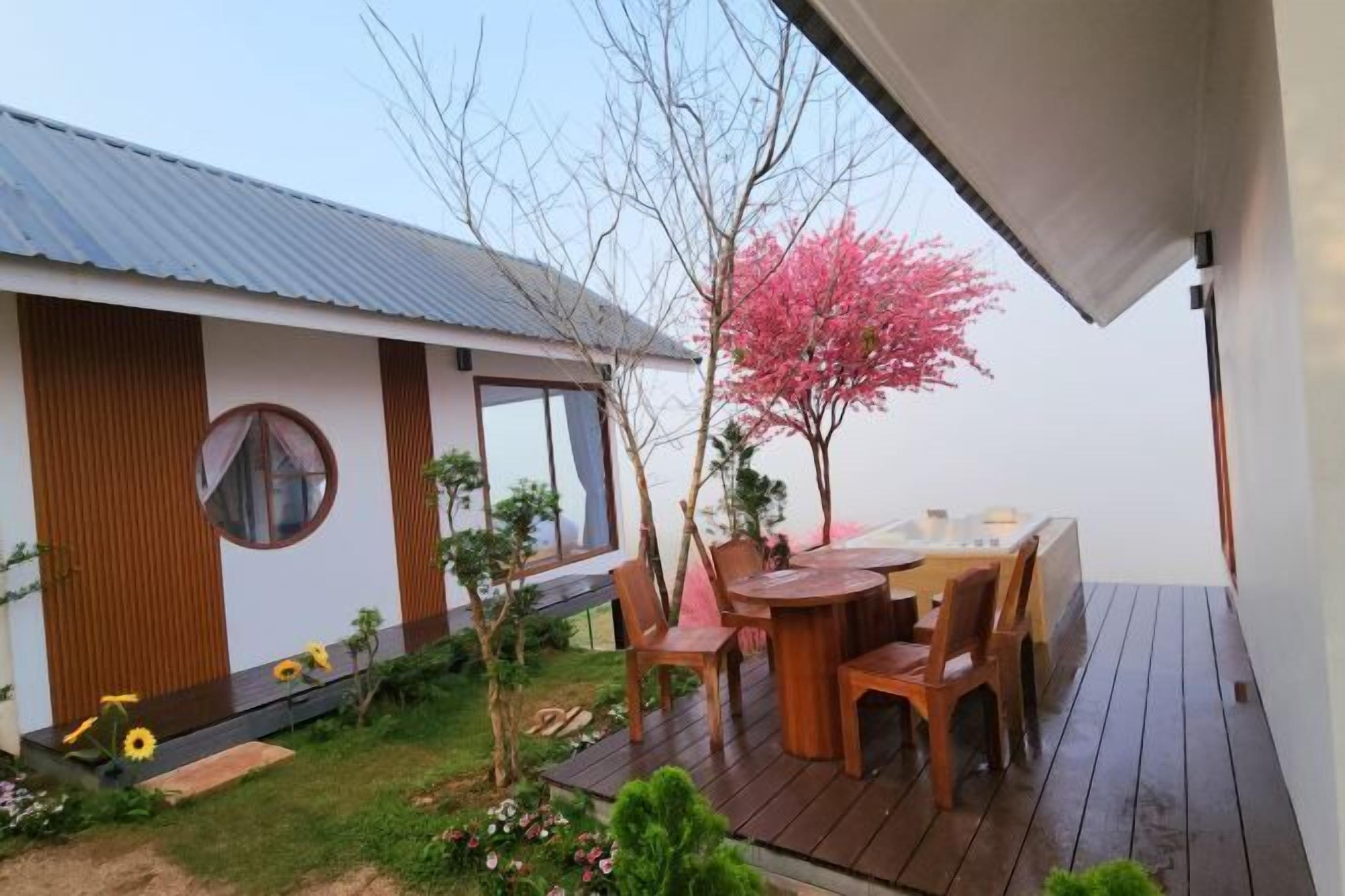A modern house with a wooden deck, a dining area with chairs, and a small garden with flowers and a tree.