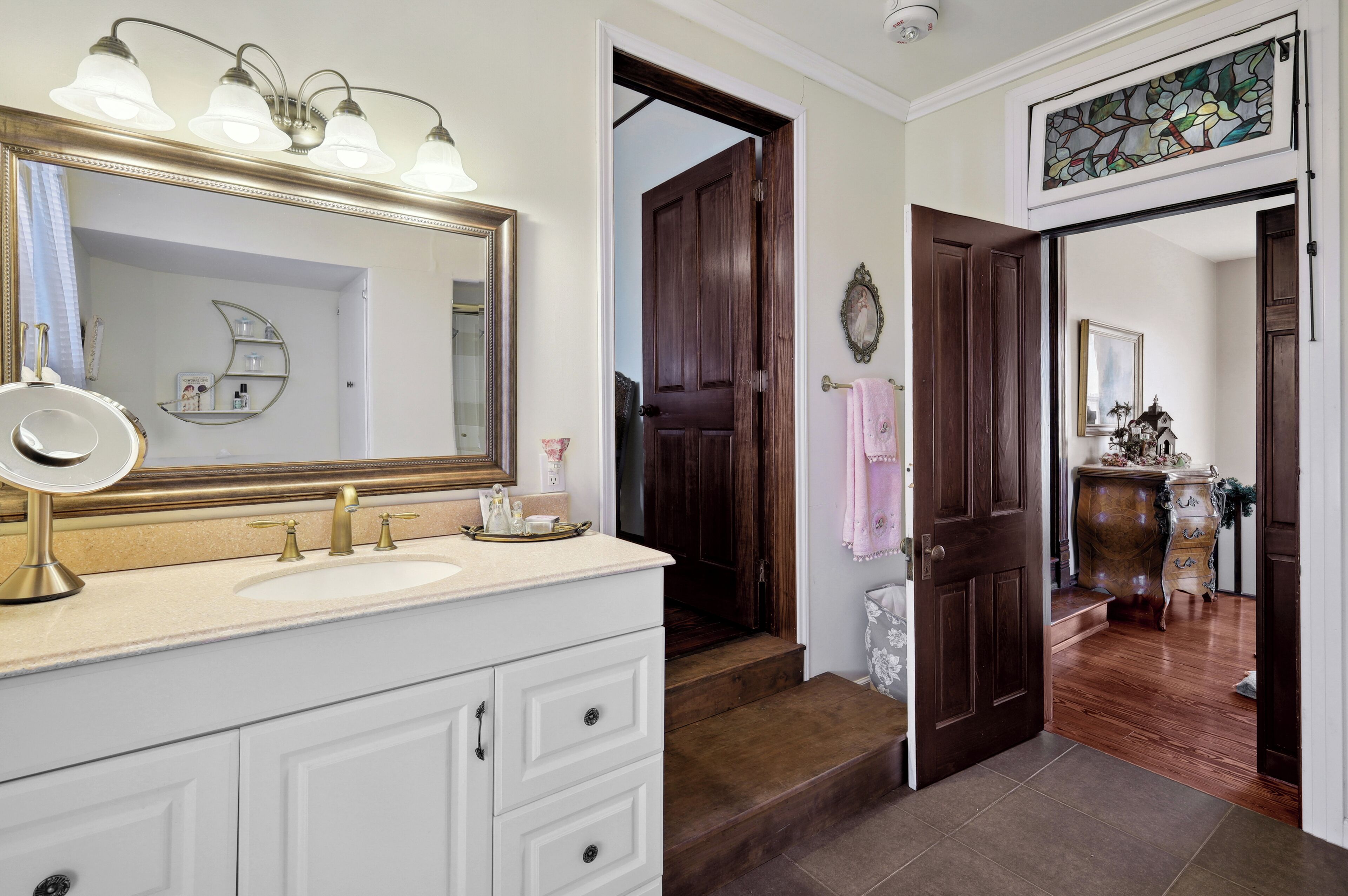 Un baño con un espejo grande, un lavamanos y un mueble de tocador. Una puerta de madera conduce a otra habitación.