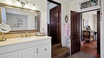 Un baño con un espejo grande, un lavamanos y un mueble de tocador. Una puerta de madera conduce a otra habitación.