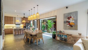 A modern kitchen with pendant lighting, a dining area with a table and chairs, and a living space with a bookshelf and a large abstract painting.