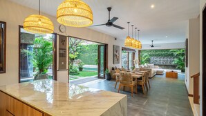 A spacious living area with a marble countertop, wicker pendant lights, ceiling fans, and a view of a lush garden.