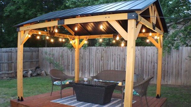 A wooden gazebo with string lights, two wicker chairs, and a table.