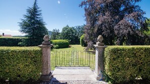 A garden entrance with a paved pathway, a metal gate, and stone pillars with sculptures. - Villa Solzago - MyHomeinComo (Tavernerio)