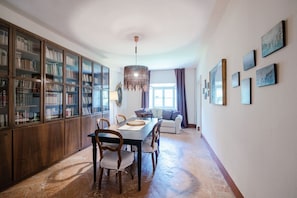 A dining area with a wooden table and chairs, a bookshelf, a hanging light fixture, and a sofa in the background.