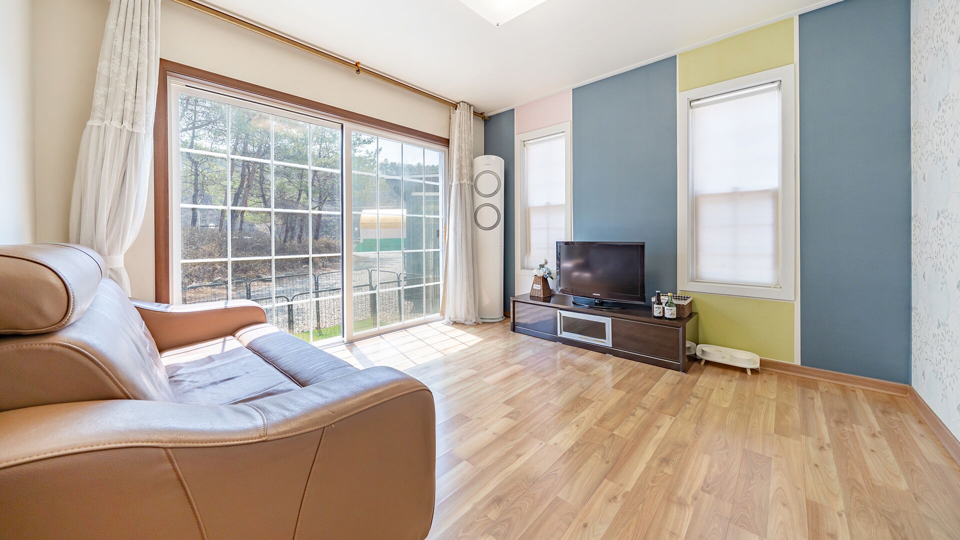 Una sala de estar con un sofá, un televisor sobre un mueble bajo y un ventanal con vistas a árboles.