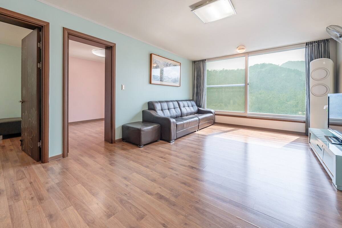 A spacious living room with a wooden floor, a leather sofa, a small ottoman, and a large window offering a view of the outdoors.