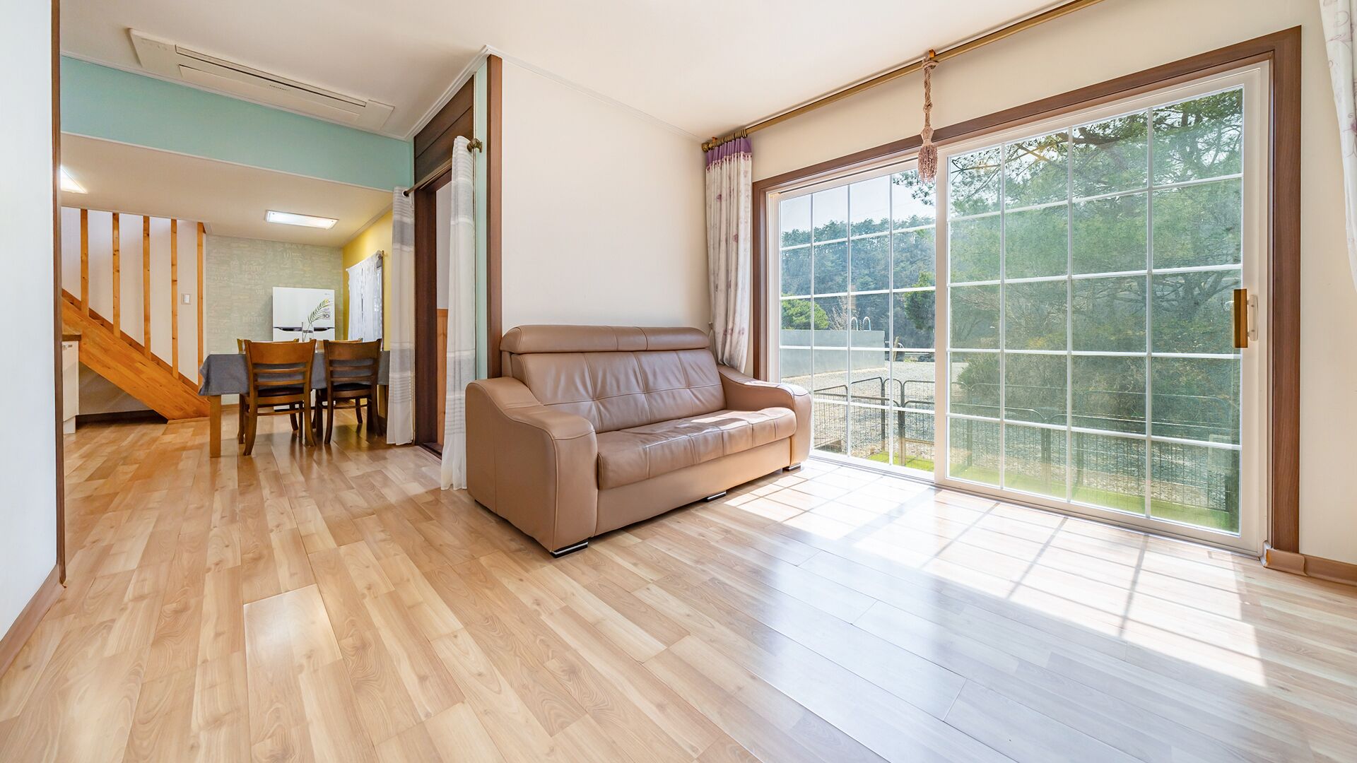 A spacious room with a wooden floor, a beige sofa, a dining area with chairs, and a large sliding glass door leading to a balcony.