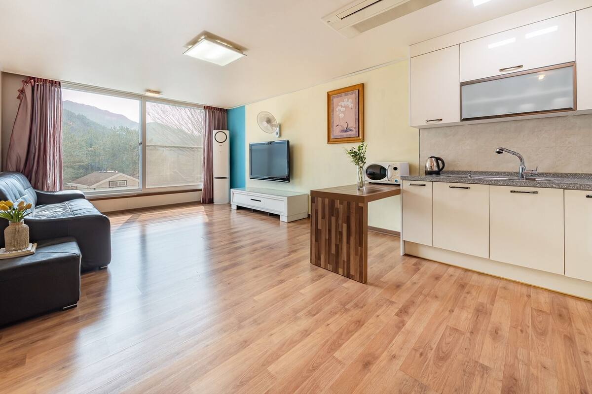 A modern living room with a sofa, a small wooden table, and a TV. There is a kitchen area with white cabinets and a countertop.
