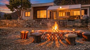 A fire pit with logs, surrounded by stone seating, in front of a modern house with large windows and a stone wall. - Yeongwol Nature & Retreat (Yeongwol)