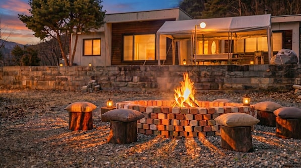 A fire pit with logs, surrounded by stone seating, in front of a modern house with large windows and a stone wall.