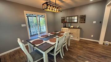 Un coin repas avec une table en bois, des chaises, un lustre et une vue sur l’extérieur, visible à travers une porte vitrée.