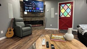 A living room with a stone fireplace, a grey armchair, a guitar, a coffee table with a book and remotes, and a red door with a stained glass window.