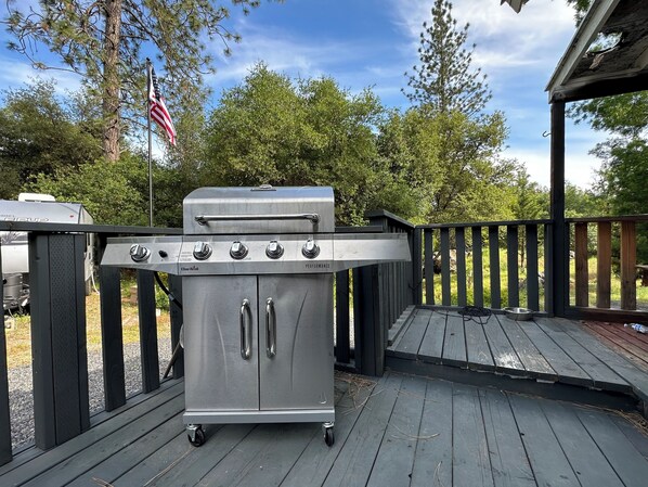 Una parrilla de gas portátil con cuatro quemadores y un quemador lateral, montada sobre ruedas, en una terraza de madera con barandilla.