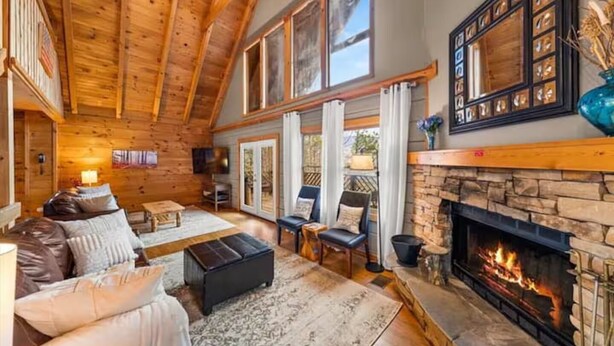 A cozy log cabin interior with a stone fireplace, wooden ceiling beams, and a large window overlooking a garden.