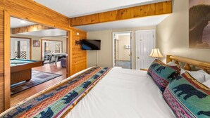 A bedroom with a large bed, a wooden ceiling, a TV mounted on the wall, and a pool table in the adjacent room.