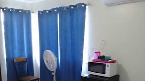Une pièce comprenant un lit, une chaise, un ventilateur, un four à micro-ondes et une petite table sur laquelle se trouvent divers objets.
