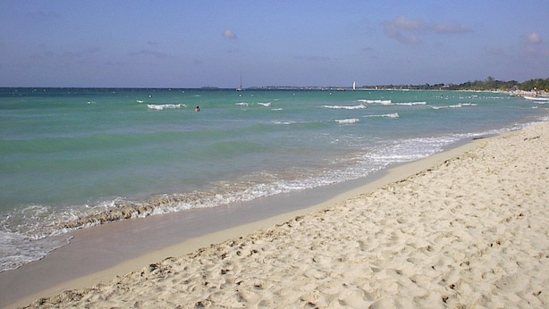 Une plage à l’eau turquoise et limpide, avec un rivage sablonneux et une vue lointaine sur des bateaux et des bâtiments.