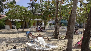 Une plage avec des chaises longues blanches, des palmiers et un bâtiment en arrière-plan.