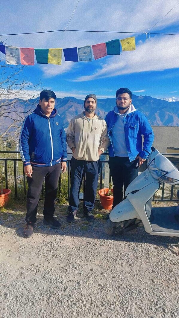Three individuals standing outdoors with a mountainous backdrop, a scooter, and a string of colorful flags.