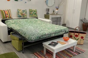 A compact room with a bed, a small table, a chair, a cabinet, and a potted plant.