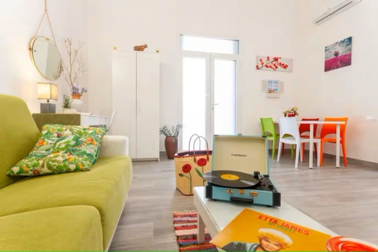 Een moderne woonkamer met een bank, een salontafel met een vinylplaat, een eethoek met stoelen, en een deur naar een andere kamer.