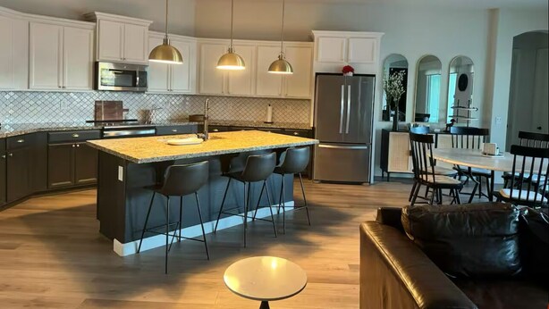 Una cocina moderna con una isla central, taburetes de bar, iluminación colgante y un comedor con sillas y una mesa.