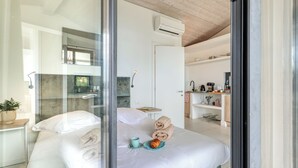 Ein modernes Hotelzimmer mit Glastrennwand, einem Bett mit Handtüchern und einem Croissant, einem kleinen Tisch mit einer Tasse sowie einer Küchenzeile im Hintergrund.