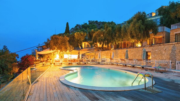 A wooden deck with a swimming pool, lounge chairs, and a stone wall.