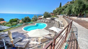 A pool area with lounge chairs and umbrellas, surrounded by greenery and overlooking a body of water. - Boutique Pool Apt. Balzi Rossi - Roma 5, Ventimiglia, Italy (Ventimiglia)