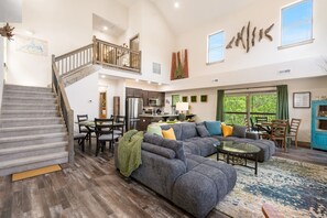 A spacious living area with a large gray sectional sofa, a dining table with chairs, a kitchen area, and a staircase leading to an upper level.
