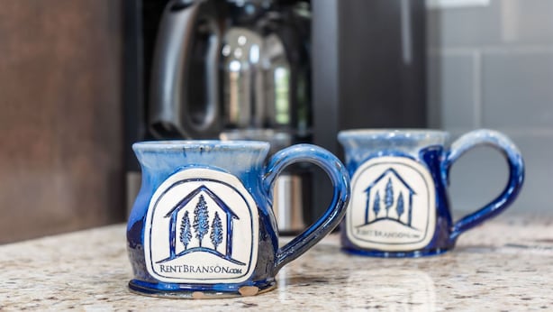 Two ceramic mugs with a house and trees logo, placed on a kitchen counter.