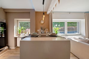 Une cuisine moderne dotée d’un îlot central, d’un coin repas avec un canapé et de grandes fenêtres offrant une vue sur la verdure.