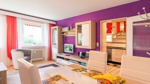 A modern room with a purple wall, a dining area with a table, a kitchen, and a window with curtains.