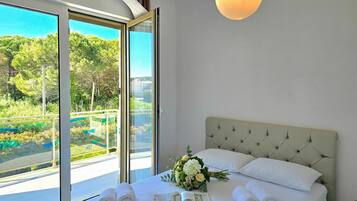 Une chambre d’hôtel avec une grande fenêtre, un lit recouvert de draps blancs, un bouquet de fleurs et un magazine posé sur le lit.