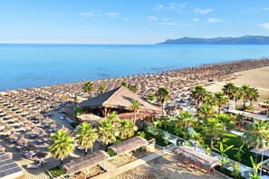 A beach with numerous thatched-roof structures and palm trees, offering a view of the sea and distant hills. - Perla Holiday Homes 276, Lalzit Bay, Albania (Lalzit Bay)