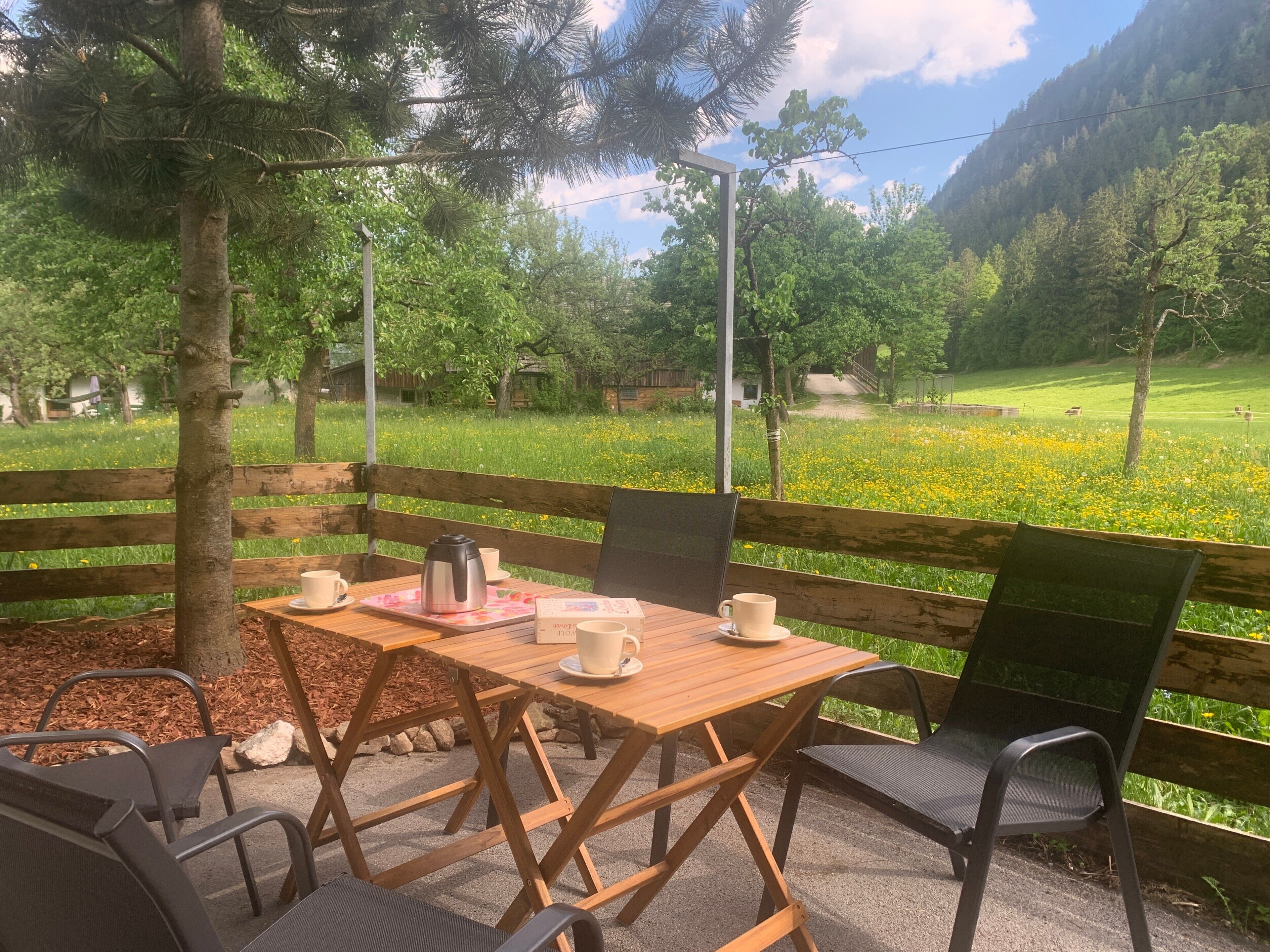 Une table en bois, entourée de chaises, avec une cafetière et des tasses, installée à l’extérieur, offrant une vue sur un champ verdoyant et des arbres.